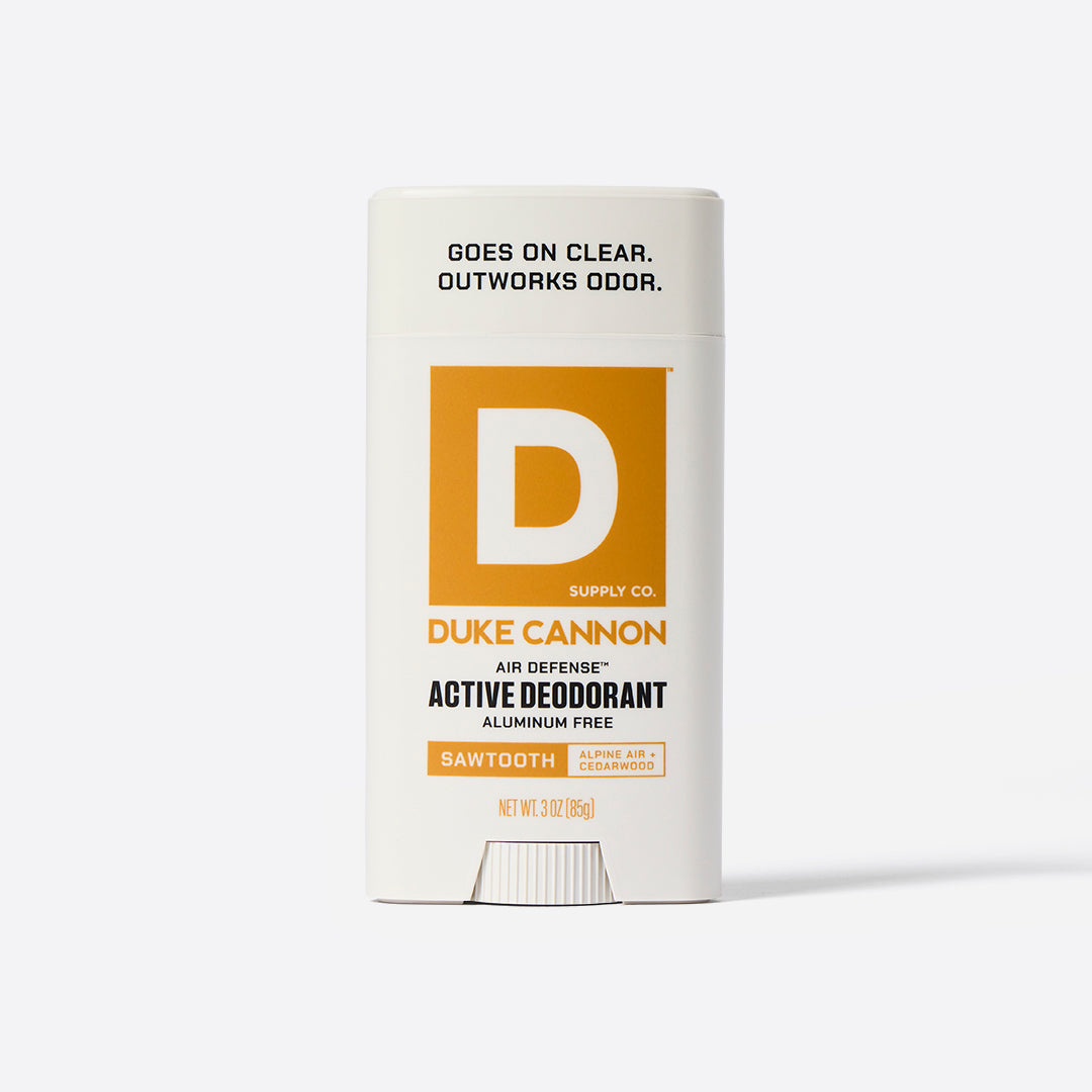 A white stick deodorant with orange and black text, labeled 'Duke Cannon Supply Co. Air Defense Active Deodorant Aluminum Free Sawtooth'. Alpine Air + Cedarwood. Says on the top, 