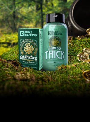 Two bottles of 'Duke Cannon' shamrock products on a forest floor with gold coins.